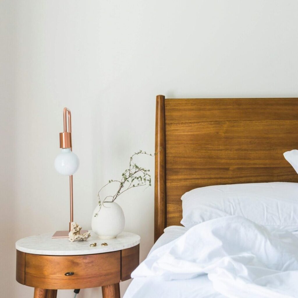 A minimalist bedroom features a wooden headboard, white bedding, and a stylish side table lamp.